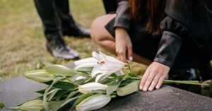 A woman in black attire mourns beside a grave, placing white lilies on the headstone in a cemetery setting.